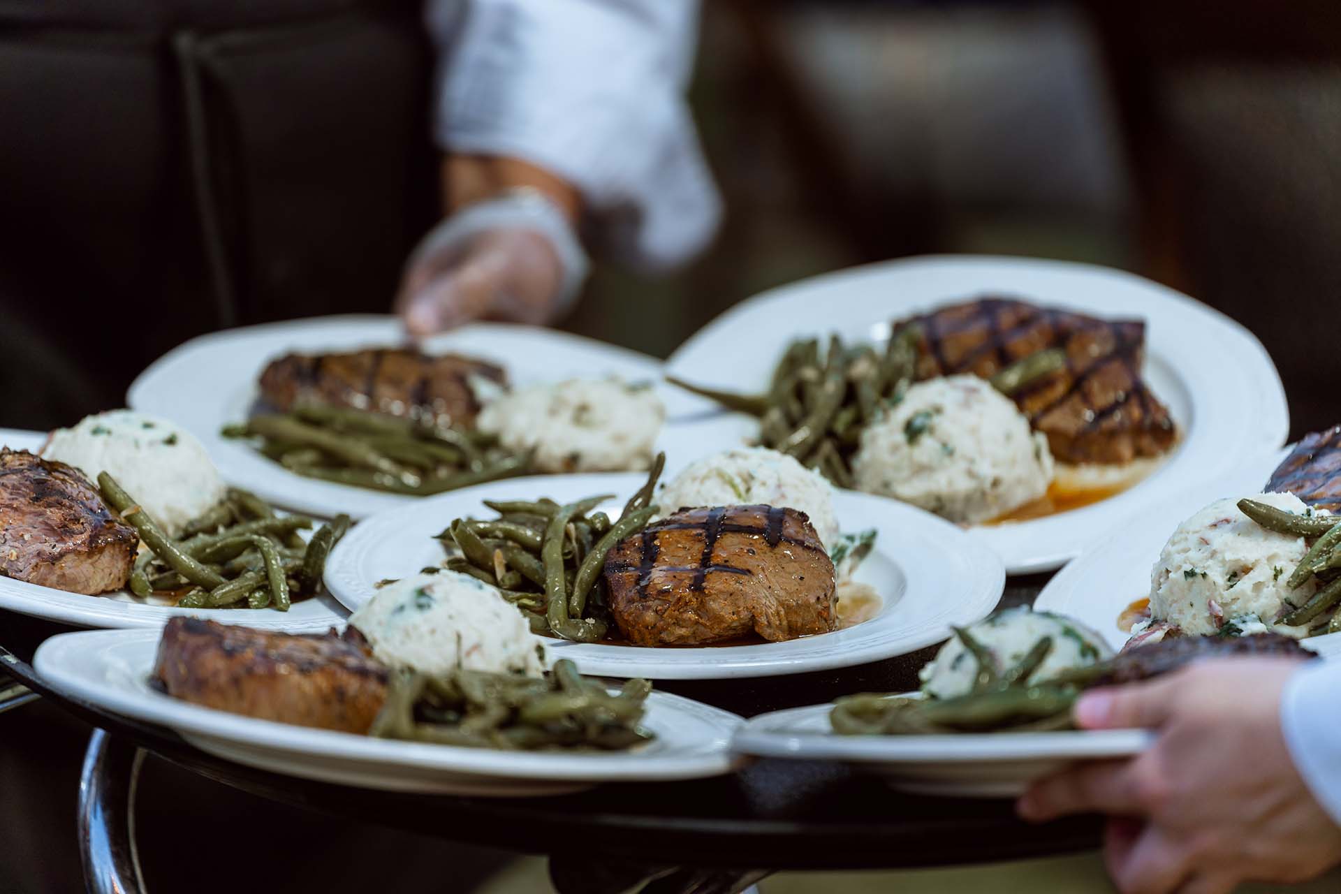 A catering staff member carries a large platter filled with dinner plates, serving up thick steaks with defined grill marks, mashed potatoes, and green beans.
