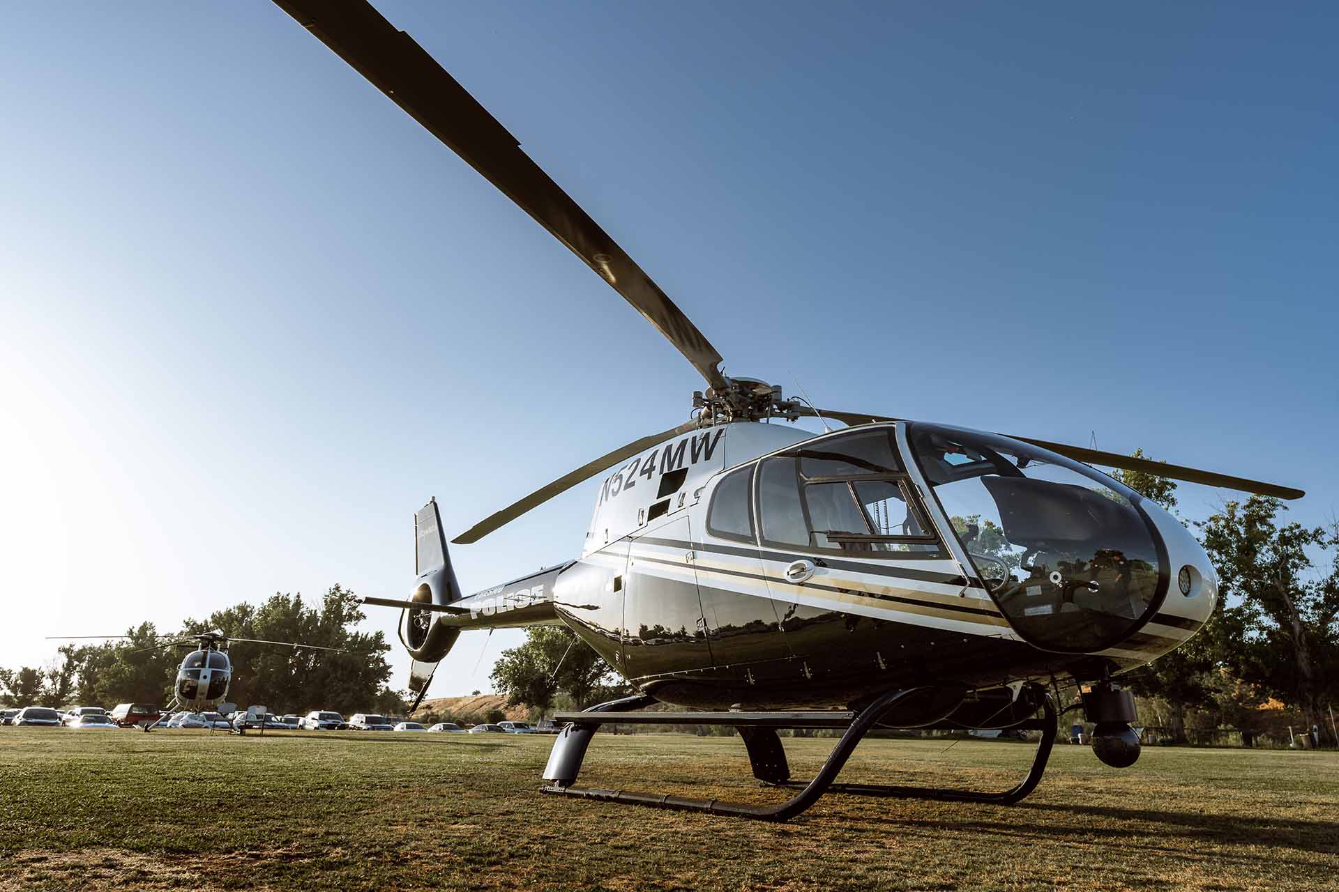 Two Fresno Police helicopters parked in a neatly mown field. The helicopters feature sleek black and silver designs with striking black and gold diagonal stripes running across the middle on the sides.