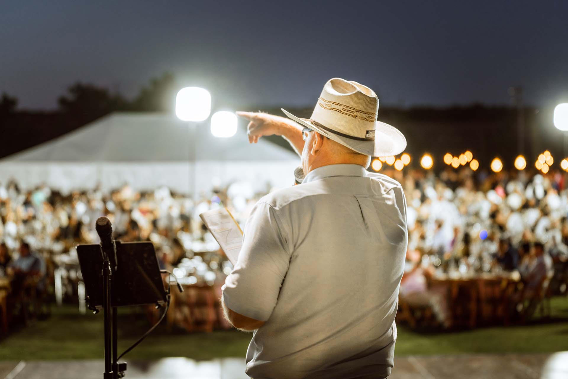 A man wearing a short-sleeved collared shirt, a cowboy hat, and glasses stands on a stage at June Jam 2023, holding a microphone and gesturing towards a crowd. The audience, seated at dining tables, fills the outdoor venue under the night sky. In the backdrop, a large event tent is illuminated with glowing lights, enhancing the festive atmosphere.